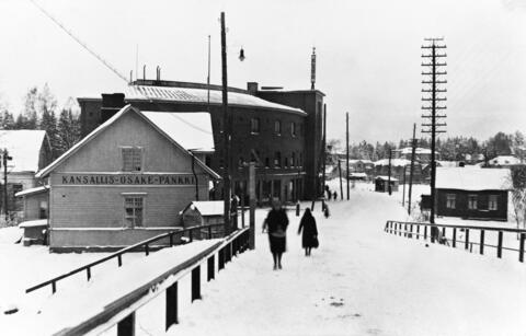 Ihmisiä talvimaisemassa Malmin Kirkonkyläntiellä. Vasemmalla Kansallisosakepankin ja Elannon rakennukset, oikealla ja taustalla pienempiä asuinrakennuksia.