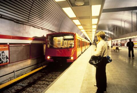 Metrojuna on juuri pysähtymässä laiturille Hakaniemessä. Etualalla metroa odottaa kahta laukkua kantava, villapaitaan pukeutunut nuori poika.