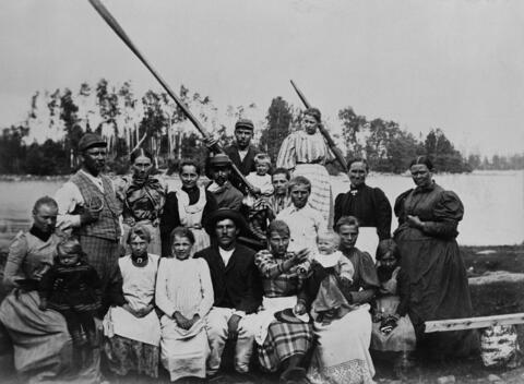 Joukko työläisiä, miehiä, naisia ja muutama lapsi, kokoontuneena veneeseen meren rantaan.