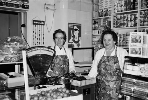 Två kvinnor arbetande i en köttbutik. Butikens väggar omges av hyllor fyllda med konserver. På disken finns produkter och en stor köttvåg. 
