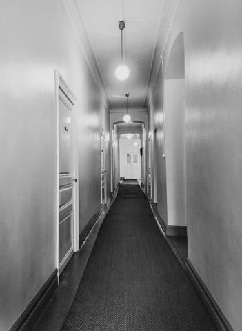 A black and white photo of an office corridor. Wooden doors on the left. Round lamps are hanging from the ceiling.