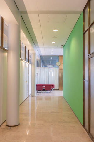A photo of an office corridor. There are columns and doors on the left, and a green wall and a copper-framed glass wall on the right.