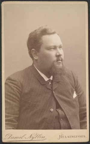 En svartvit bild på en kraftigt byggd man med mustasch och skägg. Han är klädd i kritstrecksrandig kostym.
