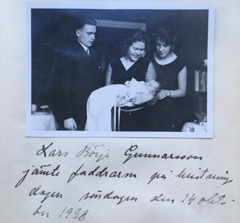 En kvinna håller ett dopklätt barn i famnen. Runt henne står en man och en kvinna, alla tittar på barnet.