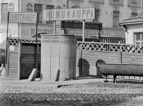 En oval pissoar av gjutjärn framför ett staket. Ovanför staketet finns skyltar med texten ROMUKAUPPA och SKROTHANDEL. I bakgrunden finns en stenbyggnad med en butik för herrkläder och en skoaffär. I förgrunden finns kullerstensbeläggning.