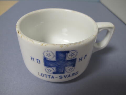 Valkoinen kahvikuppi jossa kuvituksena sininen hakaristi. Alla teksti Lotta Svärd ja sivuilla HD ja NP