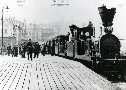 Ånglok på järnvägsstationen. På stationens träperrong står en folkskara. 