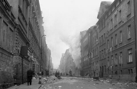 Meritullinkatu on on roskien ja rakennuksista irronneiden palasten peitossa. Talojen ikkunat ovat auki ja ikkunoiden alareunoissa on pitkiä jääpuikkoja.a. Yhdestä rakennuksesta tulee voimakkaasti savua. Kadulla liikkuu useita ihmisiä.