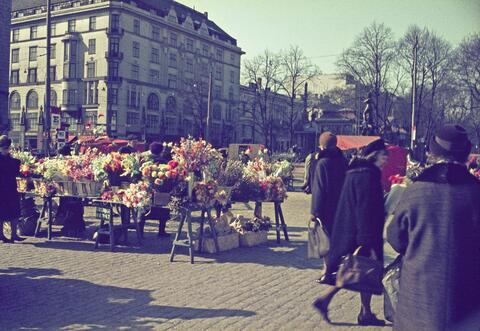 Färggranna blommor och påskprydnader på torget. Människor promenerar i förgrunden.