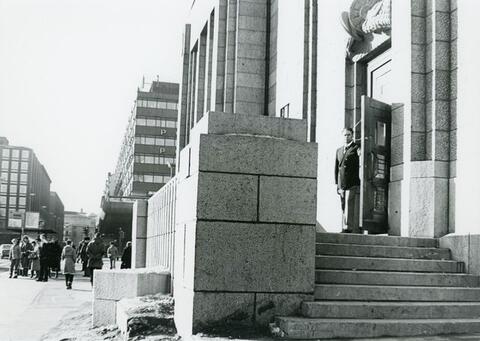 Vahtimestari seisoo ravintolan ovella mustavalkoisessa valokuvassa.
