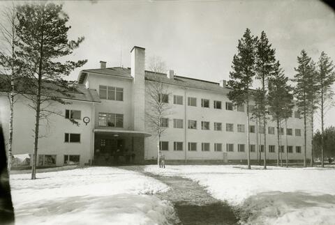 Bild av en ljus skolbyggnad i funkisstil. I förgrunden tallar och en snöig skolgård.