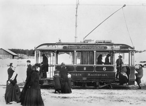 Eldriven spårvagn stannar vid hållplats, människor stiger på. Det är snö på marken. Kvinnor och barn bär långa kappor.