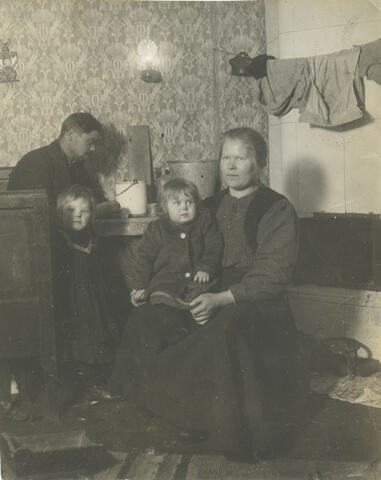 Työläisperhe (mies, nainen ja kaksi pientä tyttöä) pienessä asunnossaan n. vuoden 1910 tienoilla. Kakluunin eteen on ripustettu pyykkinaru, jolla on vaatteita kuivumassa.