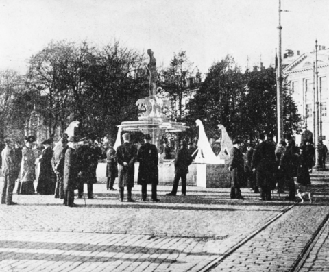 Joukko ihmisiä on kerääntynyt Kauppatorille tarkastelemaan puusta valmistettua melko aidonkokoista Ville Vallgrenin suihkukaivon mallikappaletta. Taustalla näkyy Esplanadin puistoa. Puissa ei ole lehtiä.