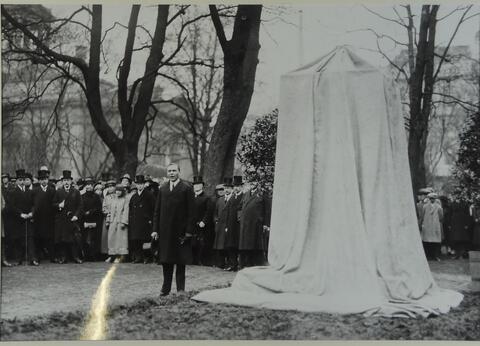 Till vänster står en man i mörk kostym, till höger står ett monument som är övertäckt med vitt tyg.