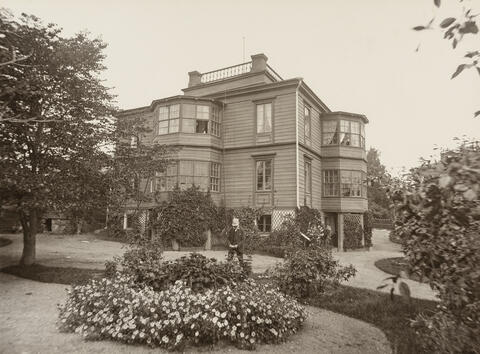 A picture of an old wooden house with three floors. Flowers and bushes in the foreground with a gardeder.