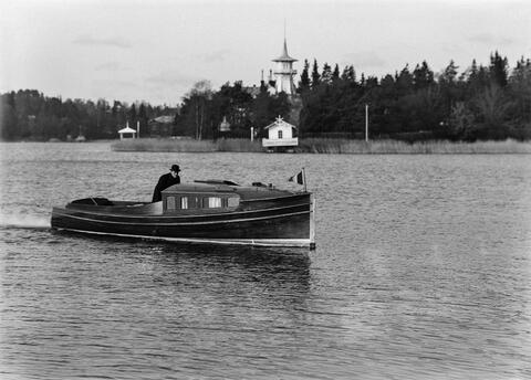 En man styr en motorbåt i trä som har en kajuta. I bakgrunden syns det som numera är statsministerns tjänstebostad och dess strand.