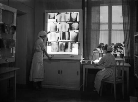 Vasemmalla seisoo nainen katsomassa valoseinään kiinnitettyjä röntgenkuvia, oikealla nainen istuu kirjoituspöydän ääressä. Hoitajien työasuun kuuluu valkoisten esiliinojen ja mekkojen ohella isolla rusetilla kiinnitetyt hilkat ja soljet paidan ylimmän napin kohdalla.