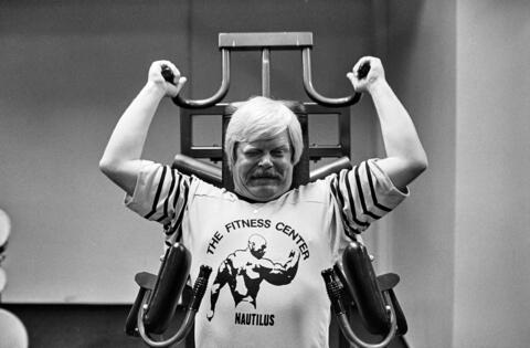 Mies kuntoilee kuntosalilaitteessa kuntosalin mainospaita päällään.