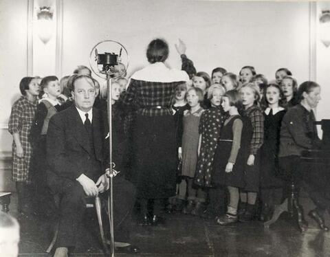 Etualalla istuu pukuun pukeutunut mies. Hänen takanaan on lapsikuoro, jota johtaa nainen selin kameraan. Oikealla istuu nainen pianoa soittamassa.