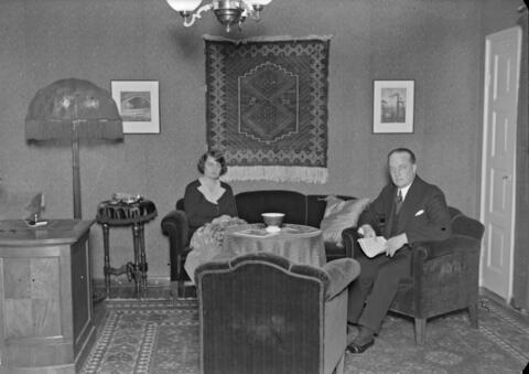 Olohuone, jossa on sohvakalusto ja ryijy seinällä. Vasemmalla istuu nainen sohvalla ja oikealla istuu mies nojatuolissa.