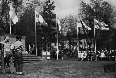 Vasemmalla esiintymiskoroke, taustalla rivi lipputankoja lippuineen. Etualalla lapsiyleisö seuraa ylioppilaslakkisten osallistujien juhlallisuuksia.