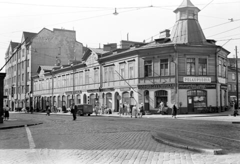 Etualalla on kaksikerroksinen puurakennus, jonka kulmassa on torni. Kulmassa on polkupyöräkor-jaamo. Kivijalassa on liikehuoneistoja, yläkerrassa asuntoja. Jalankulkijoita ja autoja. 