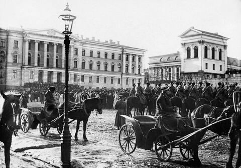Etualalla on hevosvaunuja, keskellä lukuisia miehiä ja taustalla yliopiston päärakennus. 