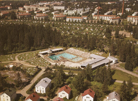 Ilmakuvassa etualalla puisia omakotitaloja, keskellä maauimala, sen takana  siirtolapuutarha ja kuvan yläreunassa harjakattoisia rapattuja kerrostaloja.