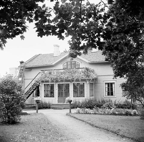 Puolitoistakerroksinen puurakennus, jossa on lasikuisti, jossa on moniruutuiset ikkunat ja katolla parveke. Etualalla on nurmikkoa ja istutuksia.