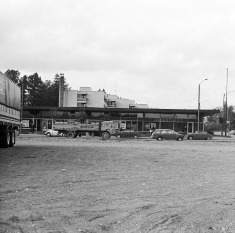 Etualalla on hiekkakenttää, keskellä on yksikerroksinen tasakattoinen ostoskeskus. Sen takana on 3–4-kerroksinen kerrostalo. Ostarin edessä on henkilöautoja ja kuorma-auto.