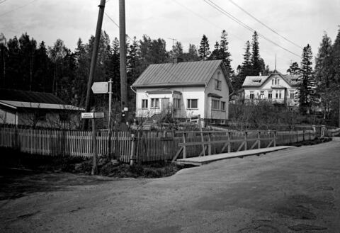 Etualalla kulkee hiekkatie. Keskellä on puuaidan reunustama omakotitalon tontti, jolla seisoo rintamamiestalo ja matala piharakennus. Taustalla on vanhempi puuhuvila. Kuvassa näkyy tieviitta, jossa lukee Eskolantie ja Unikkotie.