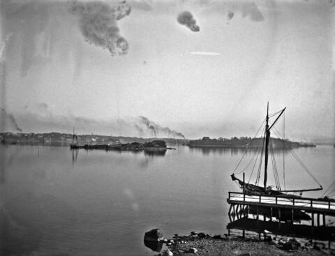 The sea and an island, with smoking factory chimneys in the background. In the foreground on the right are a quay and a sailboat. 