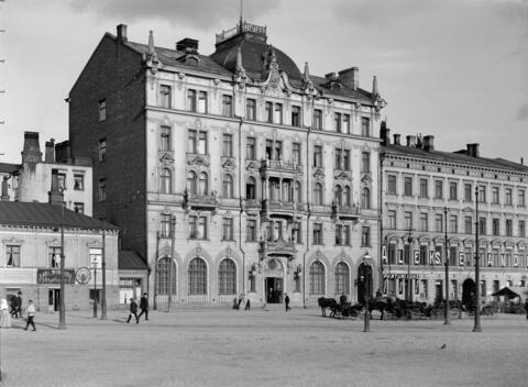 Hotelli Fennia Rautatientorilta kuvattuna. Vasemmalla olevassa puurakennuksessa on leipomo ja oikealla olevassa rakennuksessa parturinliike. Hotellin edessä on vossikkarivistö. Kadulla kulkee ihmisiä.
