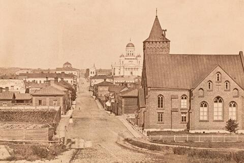 Vy mot norr längs Unionsgatan vid Tyskan kyrkan (arkitekter Harald Bosse och C.J. von Heideken 1864), Unionsgatan 1 – Bernhardsgatan 4. Till vänster i förgrunden finns en inhägnad för ved samt otydliga ljusa kvinnofigurer. I bakgrunden till vänster syns Kejserliga Alexanders-universitetet och till höger Nikolaikyrkan (1917–1959 Storkyrkan, numera Helsingfors domkyrka).