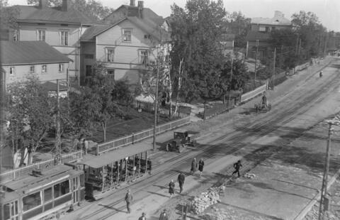 Näkymä Hämeentielle ja Hermannin suuntaan nähtynä ylhäältä alas. Kuva on otettu luultavasti vuonna 1928 valmistuneesta Hämeentie 92 kivitalosta. Hämeentietä pitkin ajaa raitiovaunu Linja 2 Hermanni - Kauppatori, joka liikennöi 1.5.1924-30.12.1928. Raitiovaunussa on avoin perävaunu. Jalankulkijoita kävelee tiellä. Hevonen vetää maitokärryä, ja ajuri seisoo kärryissä. Hämeentien varrella on puutaloja, vasemmalla Hämeentie 83 ja sen vieressä Hämeentie 81 (nykyään Hämeentie 81-83). Hämeentie 83:n sisäänkäynnin 