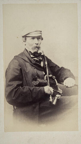 Ulkovaatteisiin pukeutunut mies istuu tuolilla iso, pitkävartinen piippu sylissään