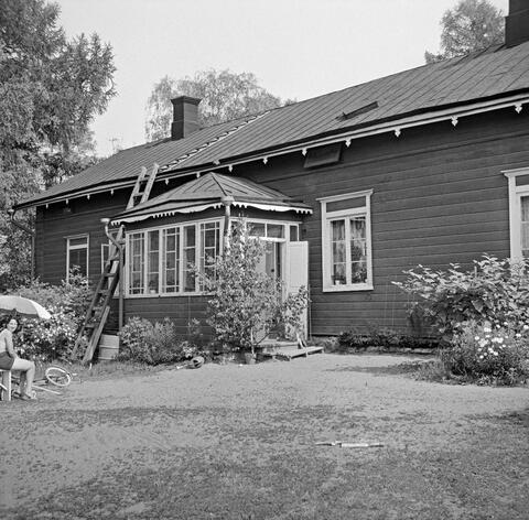 Talon pitkällä seinällä on sisäänkäynti lasikuistin kautta, rapun kulmalla koivu ämpärissä, ikkunoilla ja seinustalla kukkia. Portailla istuva lapsi ja nainen vasemmassa laidassa päivänvarjon alla katsovat kohti kuvaajaa. 