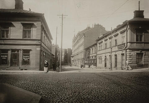 Vasemmalla Pieni Roobertinkatu 10 (- Korkeavuorenkatu 20), oikealla Pieni Roobertinkatu 7 (- Korkeavuorenkatu 22), 9, 11, 13. Liikehuoneistoissa mm. Sekalaisten Huonekalujen Kauppa ja Lähetys-Kirjakauppa, kadulla jalankulkijoita.