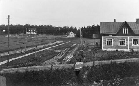 Peltoja, kapeita hiekkateitä ja kolme puurakennusta, joista yhdessä on kauppa. Sen kuistilla seisoo ihminen. 