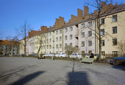 Asuinrakennukset vasemmalta oikealle Torkkelinkuja 13, 11, 9, 7, 5, 3. Äärimmäisenä vasemmalla takana häämöttää Torkkelinkuja 24. Etualalla Torkkelinpuistikko. Puistikossa on kaksi puistonpenkkiä, joista toisella istuu kaksi ihmistä. Puistikon ja asuinrakennusten väliin on pysäköity henkilöautoja. Toisen penkin takana on vaaleankeltainen Datsun 100A -henkilöauto, josta nousevalla henkilöllä on päässään keltainen lippalakki.