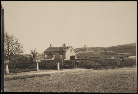 Etualalla mukulakivikatua. Keskellä on aita, jonka takana on pieni matala puurakennus pihavajoineen. Tontilla kasvaa muutama pieni lehtipuu, taustalla Tähtitorninmäen kallioita.