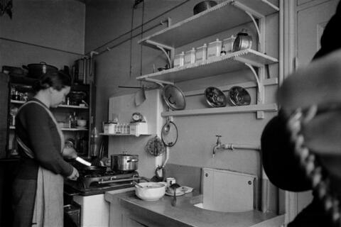 Till vänster står en kvinna och lagar mat vid en gasspis, till höger vägghyllor och ett diskbord.