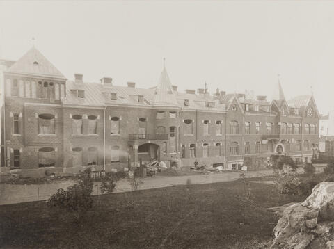 Kaksiosainen kolmikerroksinen tiilirakennus, jonka oikeanpuoleinen osa on valmis mutta vasemmanpuoleisessa ikkunoiden tilalla on säkkikangasta. Pihalla on rakennustarvikkeita.