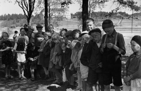 Etualalla on ryhmä lapsia kesävaatteissa. Taustalla on puita, nurmikkoa ja nelikerroksinen kivirakennus, jossa on aumakatto.