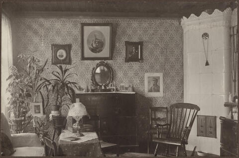 Huoneessa on kaakeliuuni, keinutuoli, kasveja sekä tauluja seinillä.