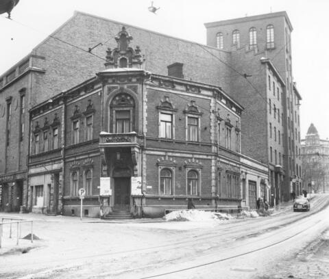 Kaksikerroksisen kulmatalon julkisivusta on poistettu tekstit ja toinen parveke. Talon takana ja sivuilla kohoaa korkean tiilirakennuksen seinät.