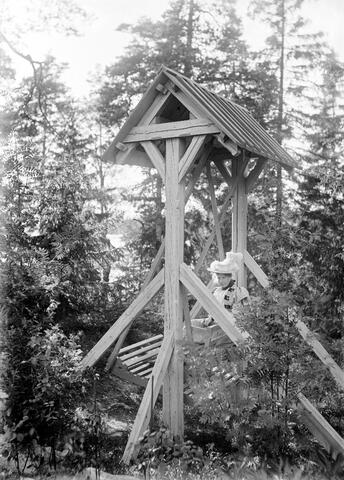 Nainen istuu katoksella varustetussa puisessa keinussa metsäisessä maisemassa. 