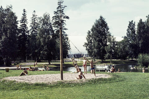 Pieni lampi, jonka ympärillä on hiekkaa ja nurmea. Poikia leikkimässä, taustalla Pirkkolan liikuntapuiston jäähalli. 