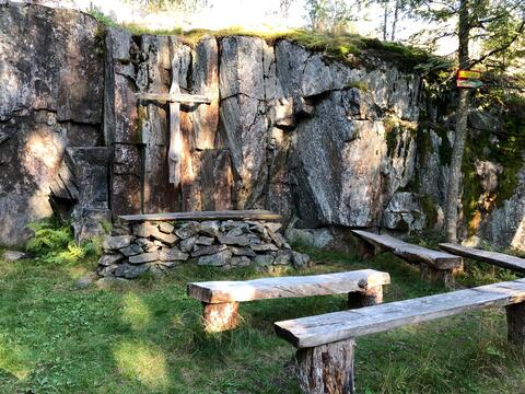 Metsäkirkko. Pystysuoraan, muutaman metrin korkuiseen kallioseinämään on kiinnitetty puinen risti. Ristin edustalla on irtokivistä rakennettu alttari ja muutama puupenkki.